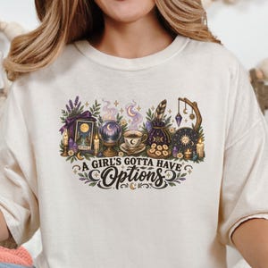 May include: An ivory crewneck sweatshirt featuring a colorful graphic print of mystical items including a tarot card, crystal ball, coffee cup, and moon phases. The text "A GIRL'S GOTTA HAVE OPTIONS" is printed in black below the illustration.