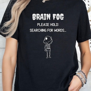 May include: A black short-sleeved t-shirt featuring white text that reads BRAIN FOG PLEASE HOLD: SEARCHING FOR WORDS... and a small white line-art illustration of a skeleton scratching its head in a thoughtful pose.