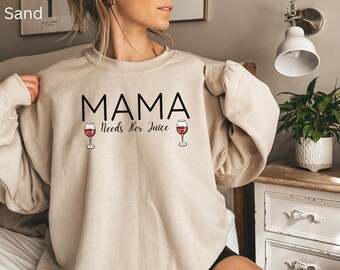 May include: Sand-colored sweatshirt with the text "MAMA Needs Her Juice" in black, accompanied by two wine glass illustrations. The sweatshirt is a neutral color and has a relaxed fit, suitable for casual wear.