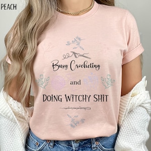 Pu&ograve; includere: Una maglietta rosa con il testo "Busy Crocheting and Doing Witchy Shit" in lettere nere. La maglietta presenta un design floreale e una piccola immagine di un uncinetto.