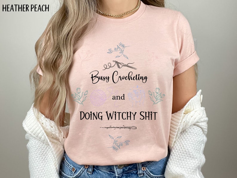 Pu&ograve; includere: Una maglietta rosa con il testo "Busy Crocheting and Doing Witchy Shit" in lettere nere. La maglietta presenta un design floreale e una piccola immagine di un uncinetto.