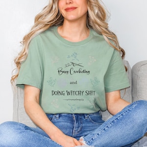 Pu&ograve; includere: Una maglietta verde salvia con un design floreale e il testo "Busy Crocheting and Doing Witchy Shit".
