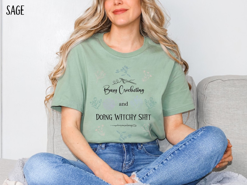 Pu&ograve; includere: Una maglietta verde salvia con un design floreale e il testo "Busy Crocheting and Doing Witchy Shit".