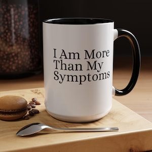 May include: White ceramic mug with a black rim and handle. The mug has black text that reads "I Am More Than My Symptoms".