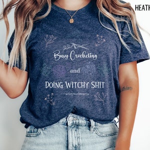 Pu&ograve; includere: Una maglietta blu navy heather con il testo "Busy Crocheting and Doing Witchy Shit" in lettere bianche. La maglietta ha un design floreale con un uncinetto e filato.