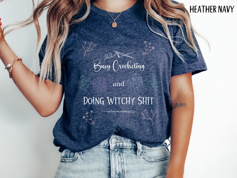 Pu&ograve; includere: Una maglietta blu navy heather con il testo "Busy Crocheting and Doing Witchy Shit" in lettere bianche. La maglietta ha un design floreale con un uncinetto e filato.