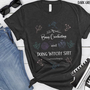 Pu&ograve; includere: Maglietta color grigio scuro melange con un design grafico bianco. La grafica presenta un gomitolo di lana, un paio di mani che tengono una luna e stelle, e il testo "Busy Crocheting and Doing Witchy Shit".