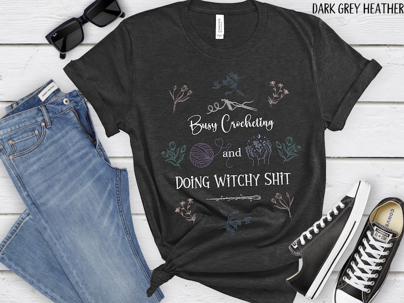 Pu&ograve; includere: Maglietta color grigio scuro melange con un design grafico bianco. La grafica presenta un gomitolo di lana, un paio di mani che tengono una luna e stelle, e il testo "Busy Crocheting and Doing Witchy Shit".