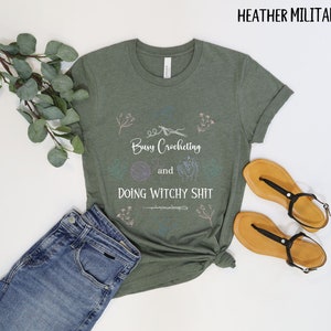 Pu&ograve; includere: Una maglietta verde con il testo "Busy Crocheting and Doing Witchy Shit" in lettere bianche. La maglietta ha un design floreale ed &egrave; su uno sfondo bianco.