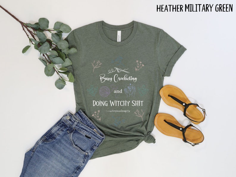 Pu&ograve; includere: Una maglietta verde con il testo "Busy Crocheting and Doing Witchy Shit" in lettere bianche. La maglietta ha un design floreale ed &egrave; su uno sfondo bianco.