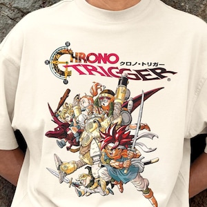 May include: Cream-colored t-shirt featuring a colorful graphic of characters from the video game Chrono Trigger. The design includes the game's logo in English and Japanese, with a group of animated figures in action poses.