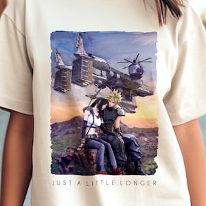May include: Cream-colored t-shirt featuring a watercolor illustration of two figures and a futuristic aircraft. The image includes the text "JUST A LITTLE LONGER" in a black font. The shirt has short sleeves and a crew neck.