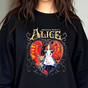 Puede incluir: Sudadera negra con un diseño gráfico de American McGee's Alice. El diseño incluye la palabra "Alice" en letras doradas, un marco en forma de corazón y una ilustración de Alicia con un vestido azul y un gato. El fondo es rojo y naranja.