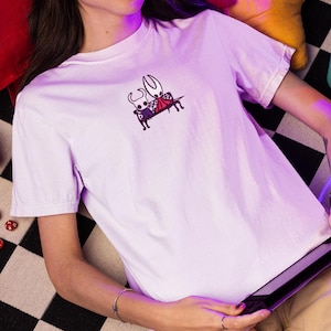 Pode incluir: Uma t-shirt de manga curta em roxo claro com um pequeno gr&aacute;fico bordado no peito. O design mostra duas personagens brancas estilizadas com chifres sentadas juntas num banco preto, uma das quais usa uma capa vermelha.