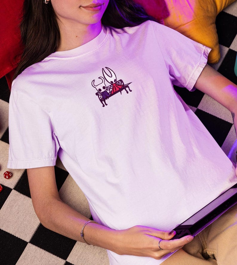Pode incluir: Uma t-shirt de manga curta em roxo claro com um pequeno gr&aacute;fico bordado no peito. O design mostra duas personagens brancas estilizadas com chifres sentadas juntas num banco preto, uma das quais usa uma capa vermelha.
