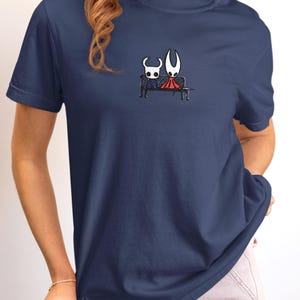 Pode incluir: Uma t-shirt azul-marinho de manga curta com um pequeno gr&aacute;fico centrado de duas personagens do jogo Hollow Knight sentadas juntas num banco preto. A personagem &agrave; esquerda &eacute; branca e azul, enquanto a da direita &eacute; branca e vermelha.