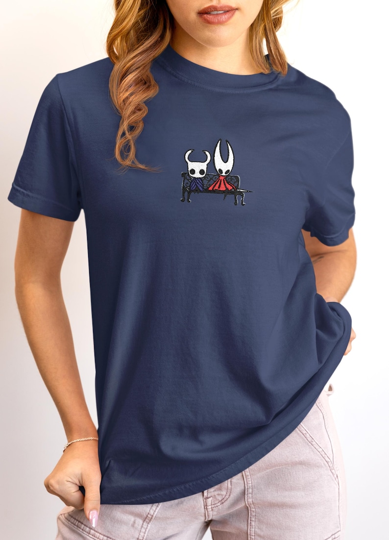 Pode incluir: Uma t-shirt azul-marinho de manga curta com um pequeno gr&aacute;fico centrado de duas personagens do jogo Hollow Knight sentadas juntas num banco preto. A personagem &agrave; esquerda &eacute; branca e azul, enquanto a da direita &eacute; branca e vermelha.