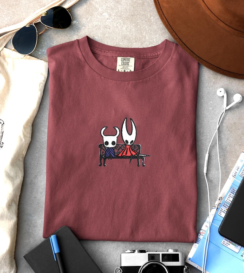 Pode incluir: Uma t-shirt de gola redonda em rosa seco com um bordado central de duas personagens brancas com chifres sentadas num banco preto. Uma personagem usa uma capa roxa e a outra uma capa vermelha. A t-shirt tem uma etiqueta Comfort Colors.