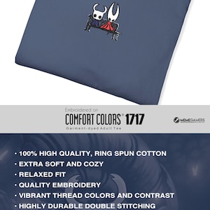 Pode incluir: Uma t-shirt azul ard&oacute;sia com um pequeno bordado centrado de duas personagens estilizadas sentadas num banco. Uma personagem &eacute; branca com detalhes pretos e a outra &eacute; vermelha com detalhes pretos. A t-shirt tem gola redonda e uma etiqueta.
