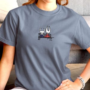 Pode incluir: Uma t-shirt de gola redonda em azul ard&oacute;sia com um pequeno gr&aacute;fico bordado no peito. O design mostra duas personagens brancas estilizadas de olhos grandes sentadas juntas num banco preto, uma com capa roxa e outra com capa vermelha.