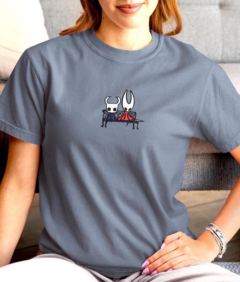 Pode incluir: Uma t-shirt de gola redonda em azul ard&oacute;sia com um pequeno gr&aacute;fico bordado no peito. O design mostra duas personagens brancas estilizadas de olhos grandes sentadas juntas num banco preto, uma com capa roxa e outra com capa vermelha.
