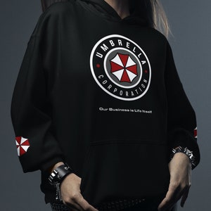 Może przedstawiać: Czarna bluza z kapturem z białym okrągłym logo przedstawiającym czerwony i biały parasol z napisem "Umbrella Corporation" i frazą "Our Business is Life Itself".