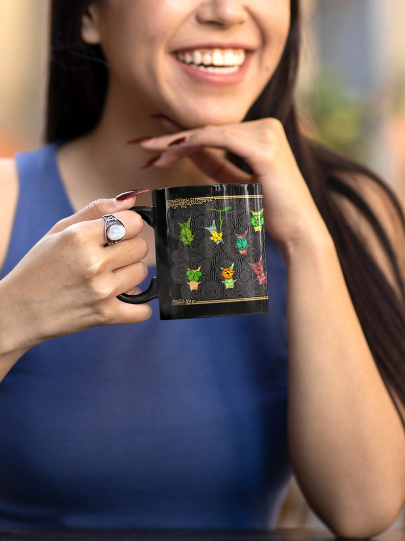 Puede incluir: Una taza de cer&aacute;mica negra con un dise&ntilde;o gr&aacute;fico colorido de caras de animales estilizadas. La taza tiene un borde dorado y una inscripci&oacute;n dorada que dice "Pick Me Up".