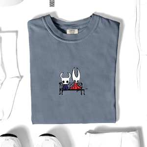 Pode incluir: Uma t-shirt de gola redonda azul ard&oacute;sia com um bordado de duas personagens brancas com chifres sentadas num banco preto. A personagem da esquerda usa uma capa azul e a da direita uma capa vermelha. A etiqueta interior diz Comfort Colors.