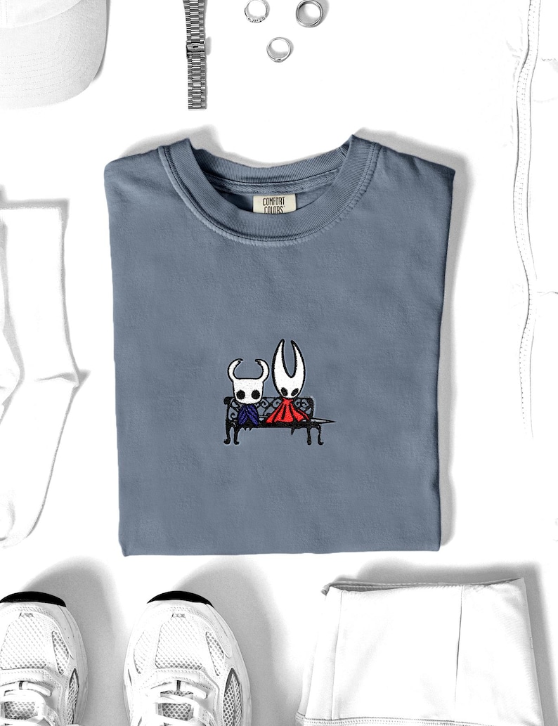 Pode incluir: Uma t-shirt de gola redonda azul ard&oacute;sia com um bordado de duas personagens brancas com chifres sentadas num banco preto. A personagem da esquerda usa uma capa azul e a da direita uma capa vermelha. A etiqueta interior diz Comfort Colors.
