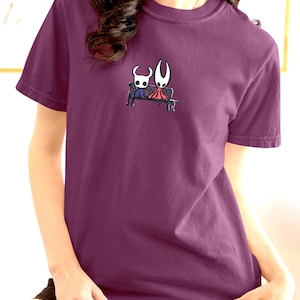 Pode incluir: Uma t-shirt de manga curta cor de ameixa com um pequeno gr&aacute;fico centrado de duas personagens estilizadas sentadas num banco preto. Uma personagem &eacute; branca com olhos escuros e a outra &eacute; branca com uma capa vermelha, em estilo de linha.