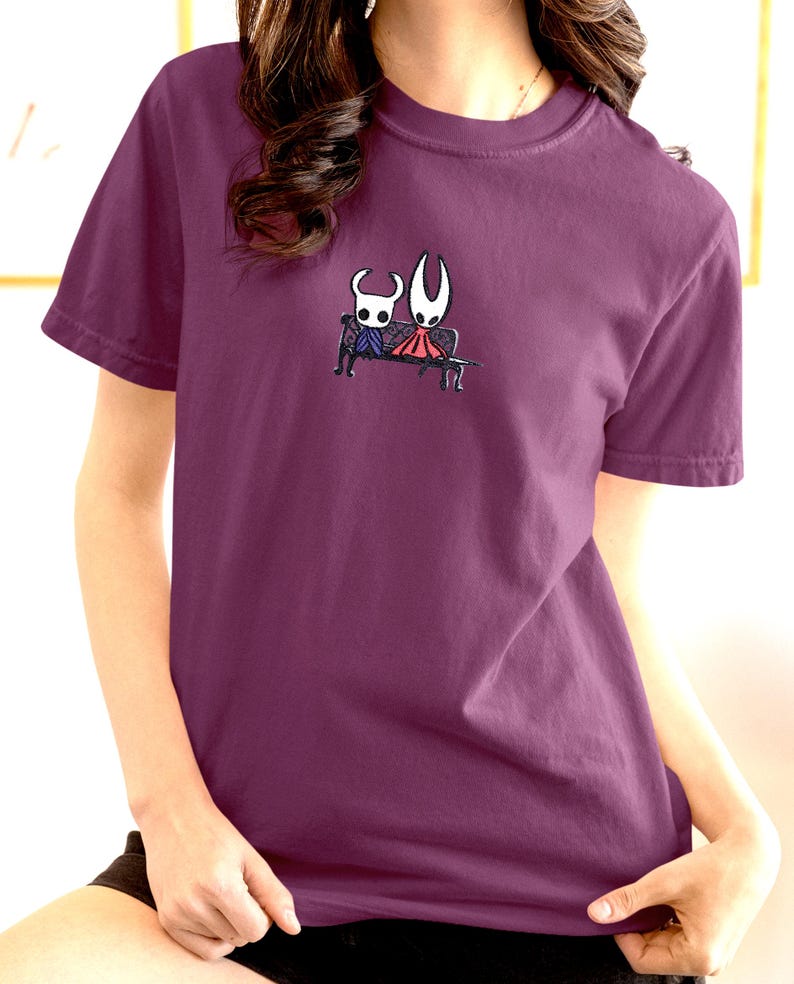 Pode incluir: Uma t-shirt de manga curta cor de ameixa com um pequeno gr&aacute;fico centrado de duas personagens estilizadas sentadas num banco preto. Uma personagem &eacute; branca com olhos escuros e a outra &eacute; branca com uma capa vermelha, em estilo de linha.