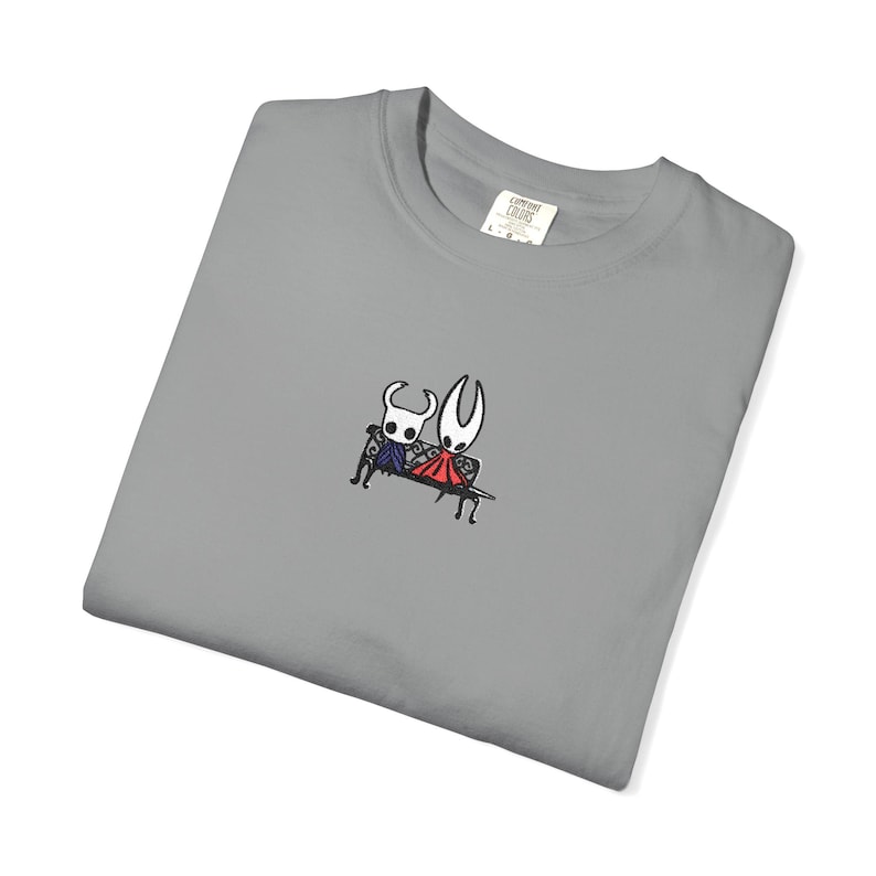 Pode incluir: Uma t-shirt cinzenta com um pequeno bordado de duas personagens brancas estilizadas sentadas num banco preto. Uma personagem usa uma capa azul-escura e a outra uma capa vermelha brilhante. A t-shirt tem uma etiqueta branca no colarinho.