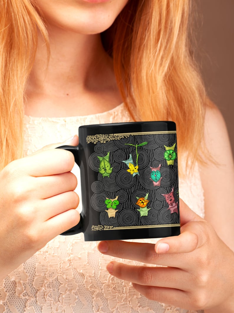 Puede incluir: Una taza de cer&aacute;mica negra con un borde dorado y un dise&ntilde;o de personajes de dibujos animados verdes, amarillos y azules sobre un fondo negro y dorado.
