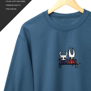 Pode incluir: Uma camisola azul de gola redonda com um bordado de duas personagens de m&aacute;scara branca sentadas num banco preto. O texto "ALSO AVAILABLE IN A SWEATSHIRT! GILDAN 18000 UNISEX SWEATSHIRT PLUSH, SOFT AND WARM PREMIUM QUALITY PRE-SHRUNK" &eacute; vis&iacute;vel.