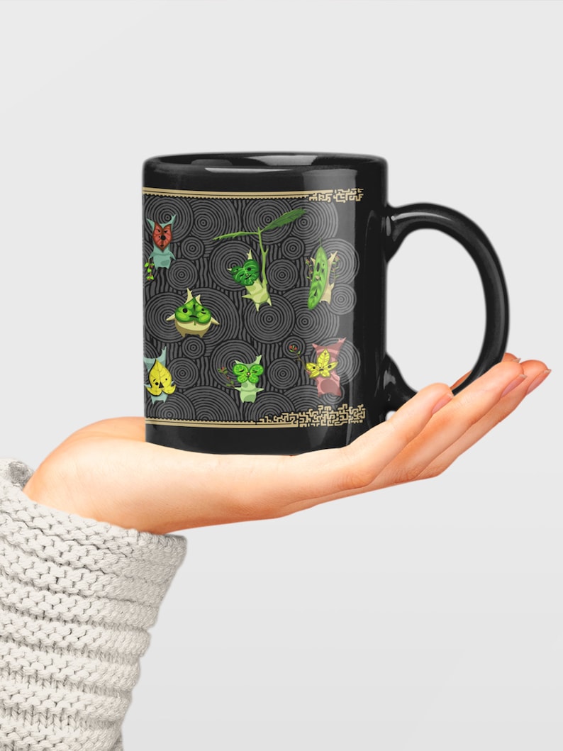 Puede incluir: Taza de cer&aacute;mica negra con un dise&ntilde;o de criaturas verdes, amarillas y rojas sobre un fondo negro y dorado. Las criaturas est&aacute;n rodeadas de c&iacute;rculos conc&eacute;ntricos.