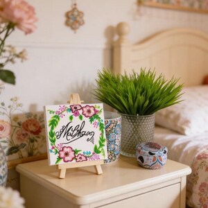 May include: A canvas painting on a wooden easel, with floral artwork and the name "Mclhan" in cursive. The painting is framed by a green and pink floral wreath. A small decorative tin and a vase with green grass are also on the bedside table.