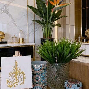 May include: An interior scene with a canvas displaying gold Arabic calligraphy on a small easel, a patterned ceramic mug, a textured glass vase with green plants, and a small decorative tin. A large potted bird of paradise plant is also present.
