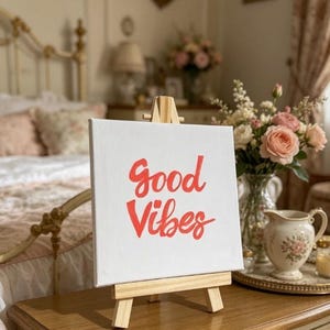 May include: A white canvas with the words "Good Vibes" in red script is displayed on a small wooden easel. The canvas is set on a wooden surface, with a vase of flowers and a decorative jug in the background.