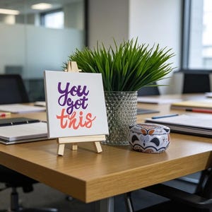 May include: A small white canvas on a wooden easel displays the text "You got this" in purple and coral. A decorative candle tin and a vase with green plants sit on a wooden table in an office setting.