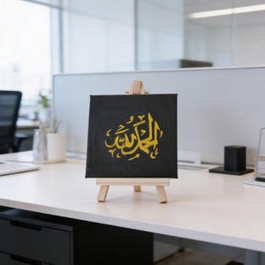 May include: A small black canvas with gold Arabic calligraphy, displayed on a wooden easel. The artwork is square and features ornate lettering. The easel is made of light-coloured wood, and the canvas is set on a white desk.