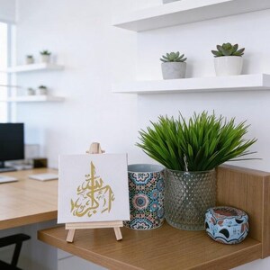 May include: A small canvas with gold Arabic calligraphy on a wooden easel sits on a wooden shelf. A patterned ceramic mug, a glass vase with green grass, and a small decorative box are also on the shelf. White shelves with potted succulents are in the background.