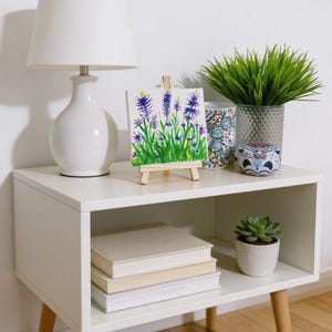 May include: A white bedside table with a lamp, artwork, and decorative items. The artwork features purple flowers on a small easel. A stack of books and a potted succulent are on the lower shelf. The table has wooden legs.