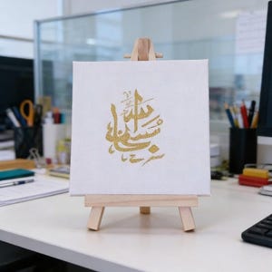 May include: A small, square white canvas with gold calligraphy displayed on a wooden easel. The calligraphy is in a flowing script. The easel is made of light-coloured wood and is positioned on a white desk. The background includes office supplies and a computer monitor.