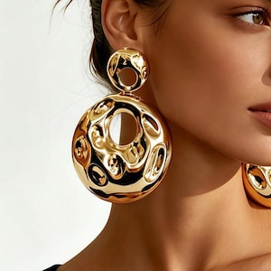 May include: A pair of large, gold-toned earrings featuring a double-circle design. Each earring consists of a smaller top circle and a significantly larger bottom circle, both characterized by a highly reflective, irregular, and hammered surface texture.