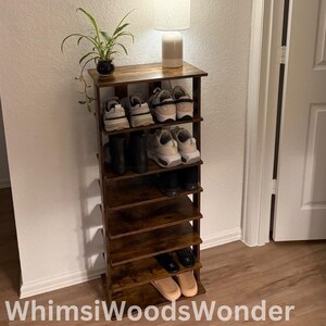 May include: A dark brown wooden shoe rack with six shelves, holding various shoes, including sneakers, boots, and sandals. A small table lamp with a white shade sits on top of the rack, and a framed picture of a tequila bottle hangs on the wall behind it.