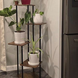 May include: A four-tier plant stand with a dark brown wood finish and black metal frame. The stand is holding four potted plants, including a large green plant with a red pot, a smaller green plant with a white pot, and two smaller green plants with white pots.