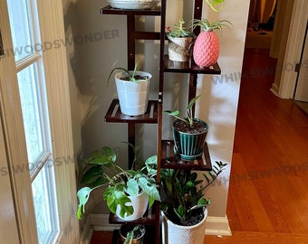 May include: A brown wooden plant stand with five shelves, holding six potted plants. The plants include a large green plant with trailing vines, a small green plant with pink flowers, and a variety of other green plants. The stand is in a room with hardwood floors and a white door.