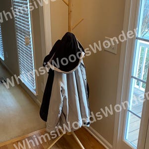 May include: A wooden coat rack with a tree-like design. A black hooded jacket and a light gray jacket hang on the rack. The rack is made of light-colored wood and stands on a wooden floor.