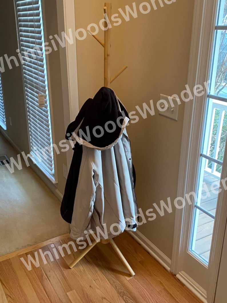 May include: A wooden coat rack with a tree-like design. A black hooded jacket and a light gray jacket hang on the rack. The rack is made of light-colored wood and stands on a wooden floor.