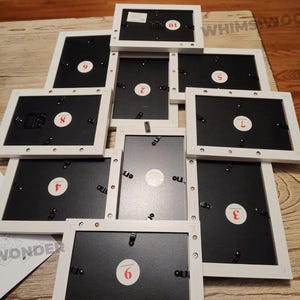 May include: Nine white picture frames with black backing and numbered labels from 1 to 9. The frames are stacked in a pyramid shape on a wooden surface.